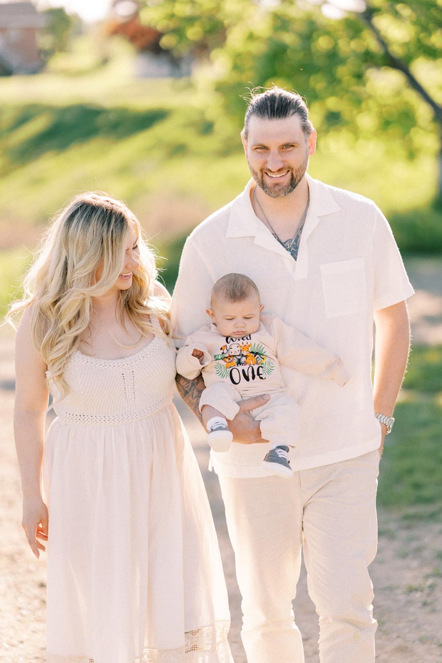 A family enjoying a sunny day outdoors, dressed in light-colored clothing, with the father holding their baby.