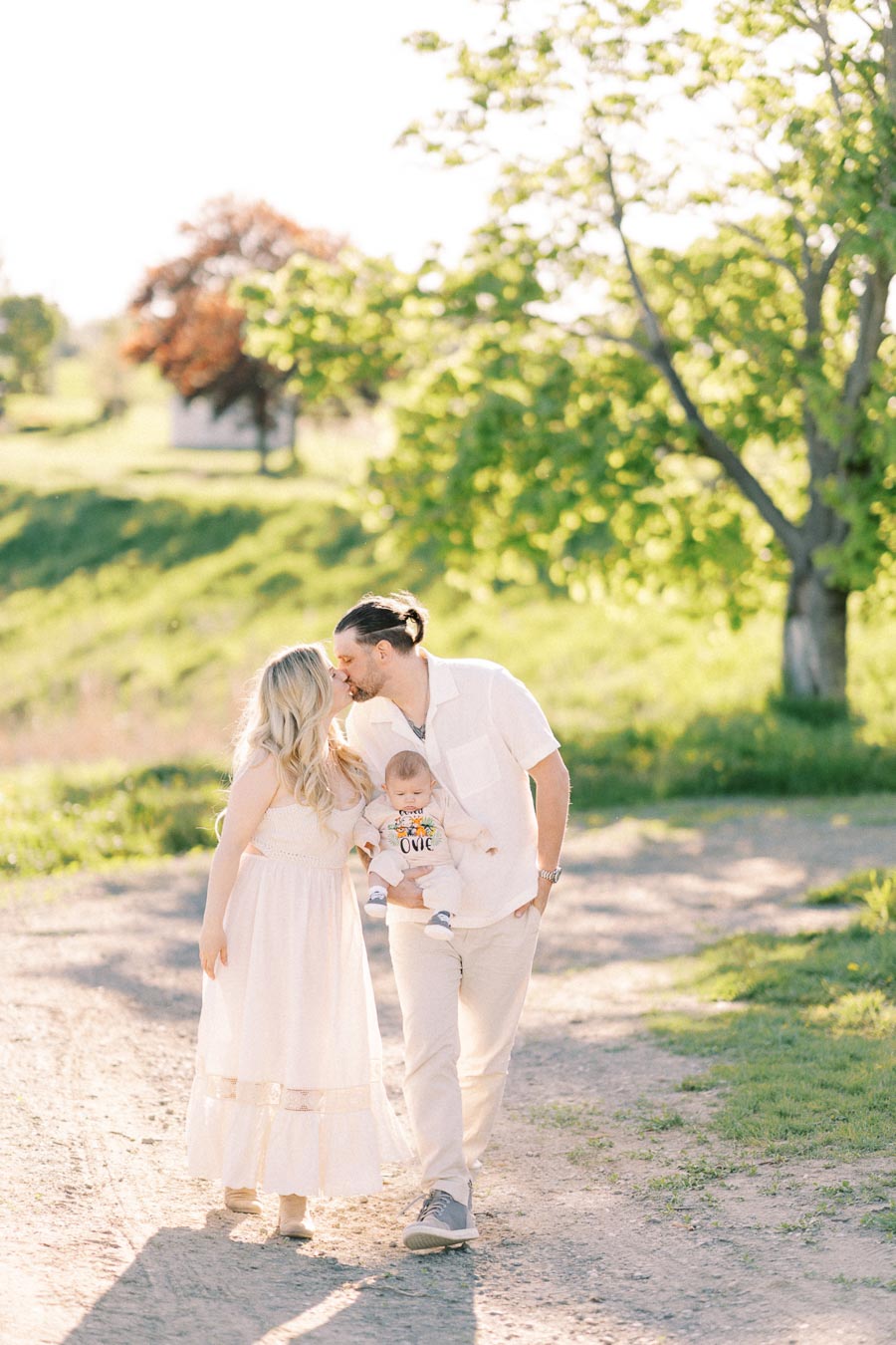 A couple shares a kiss while holding their baby as they walk along a sunlit path in a park, surrounded by lush greenery.