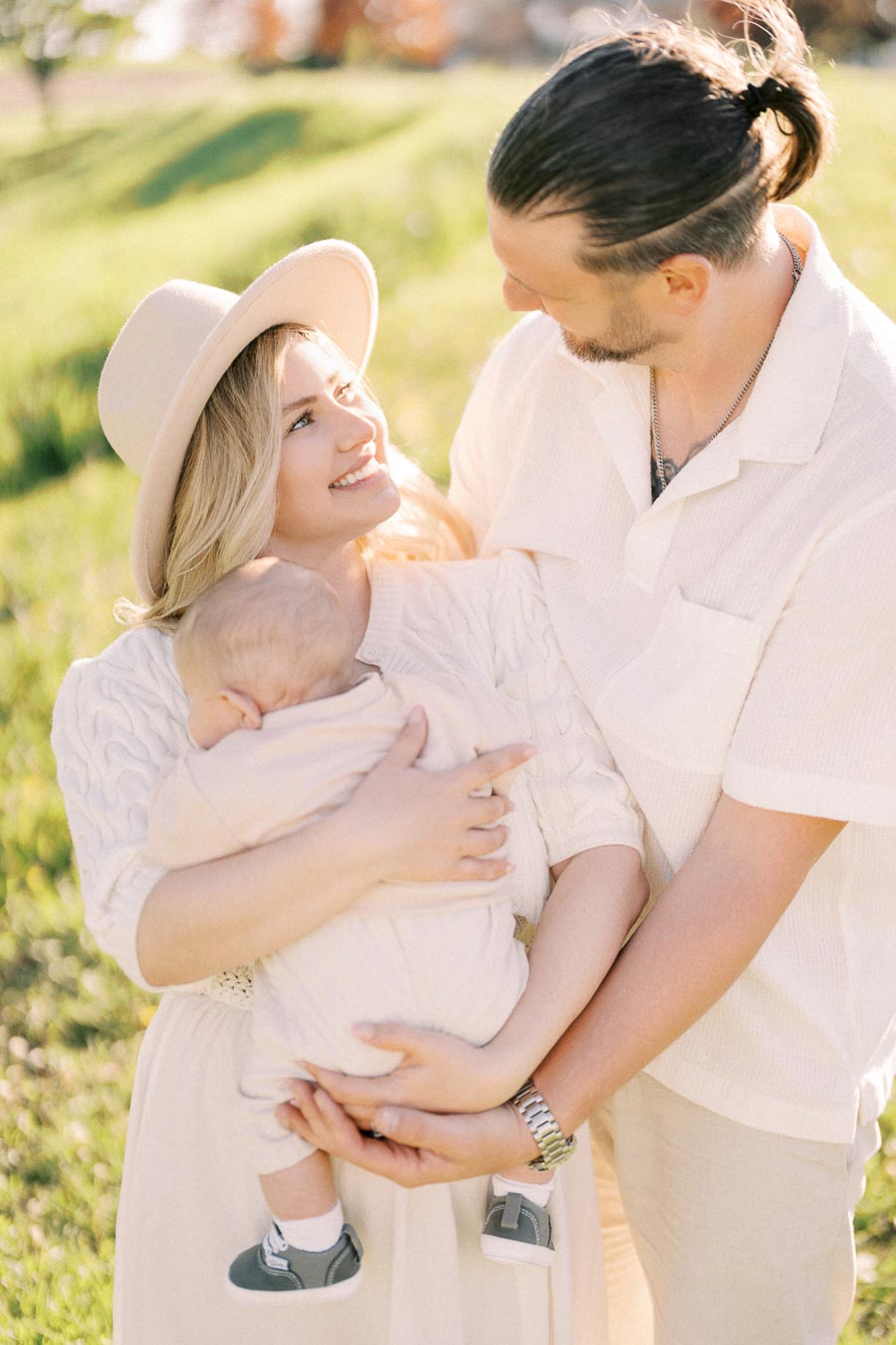 A joyful family moment captured outdoors, featuring a mother holding a baby and a father standing beside them, all dressed in light-colored clothing.