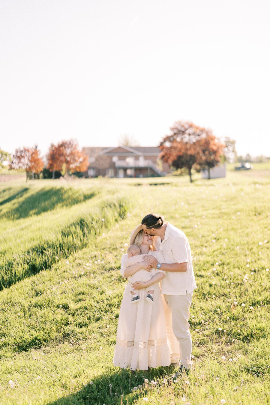 A couple embraces their baby in a sunlit field, surrounded by lush grass and distant trees, with a house visible in the background.
