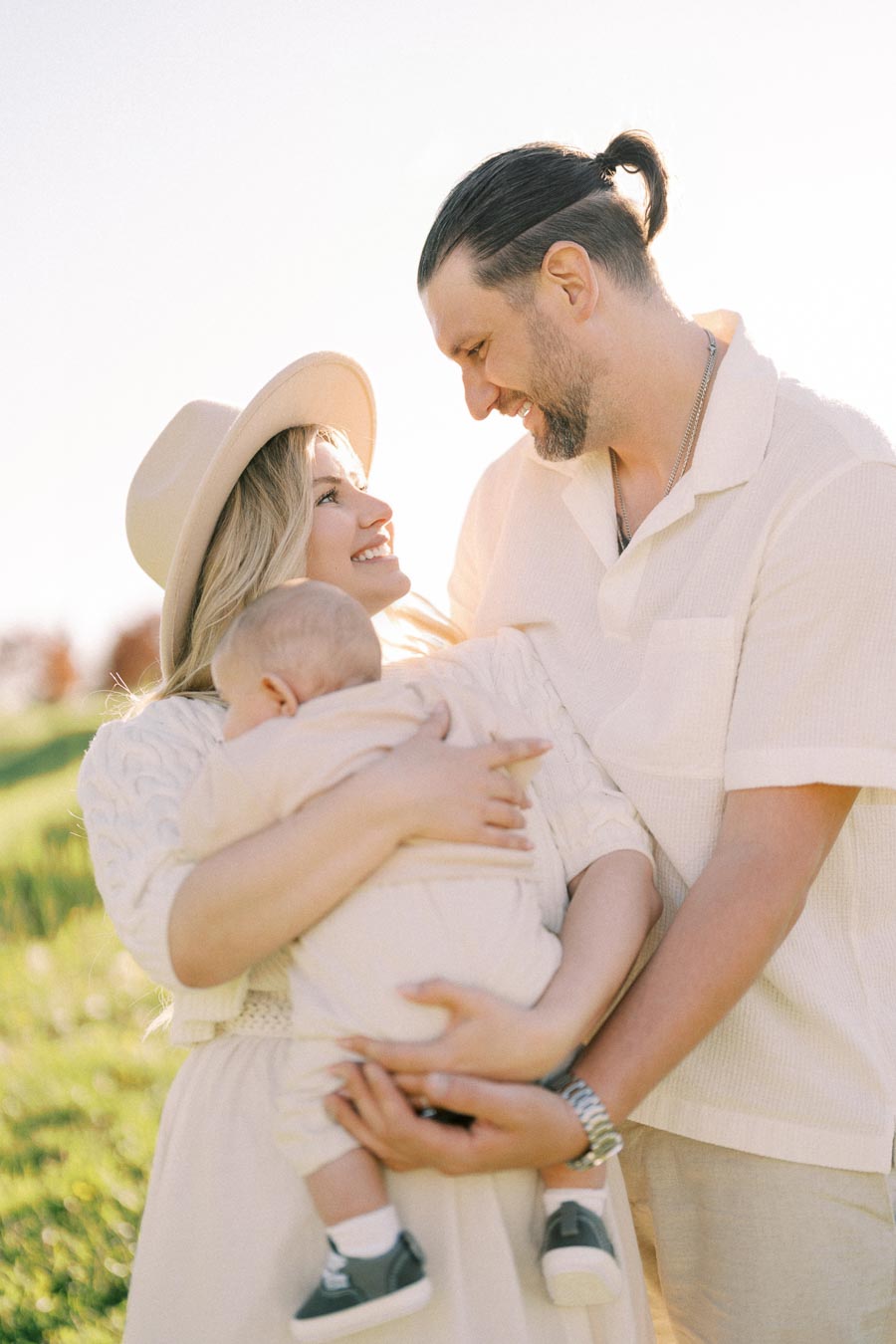 A joyful family moment captured outdoors, with a woman holding a baby and a man beside them, all smiling warmly at each other.