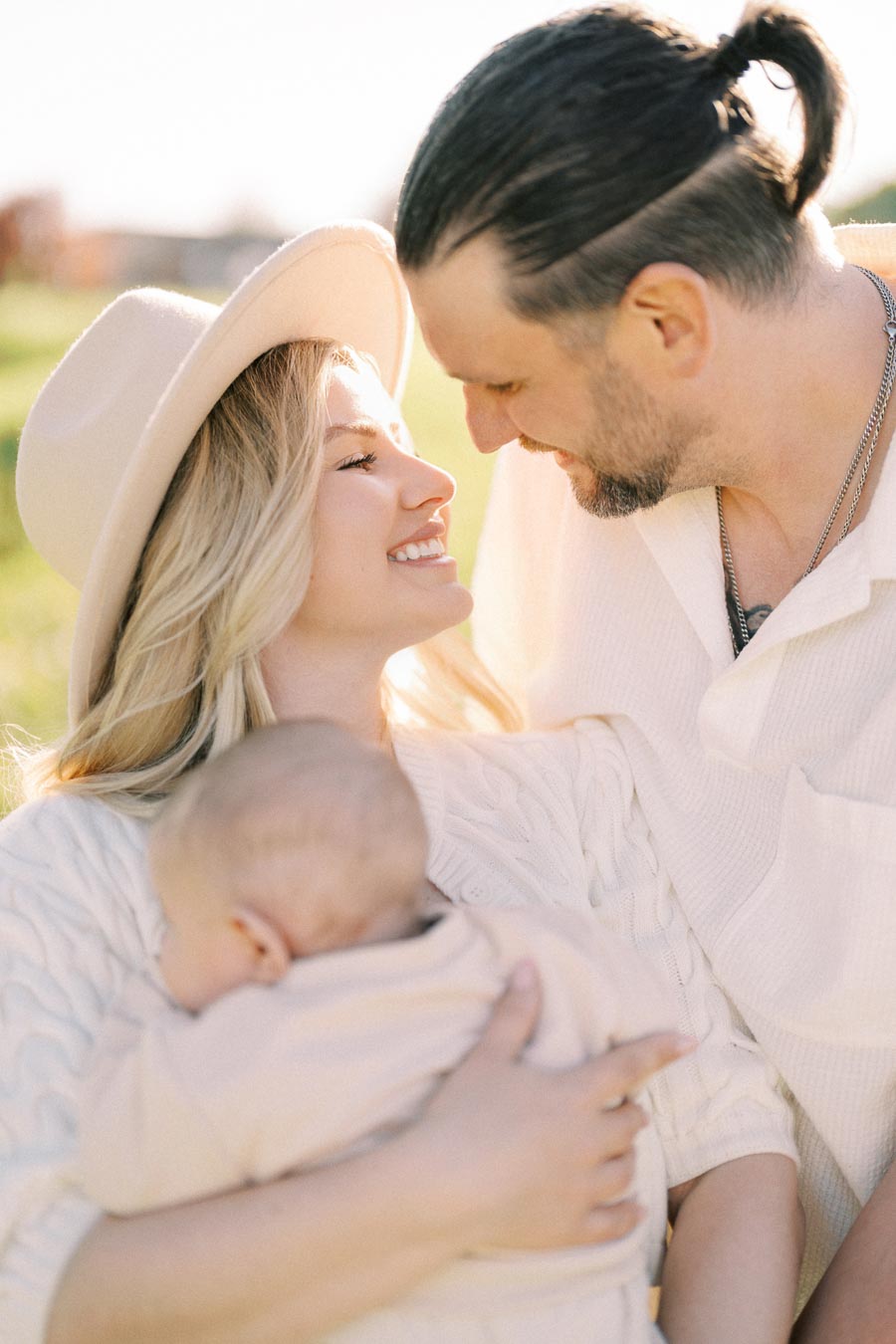 A joyful couple shares a tender moment outdoors, with the woman holding a baby and smiling at the man.