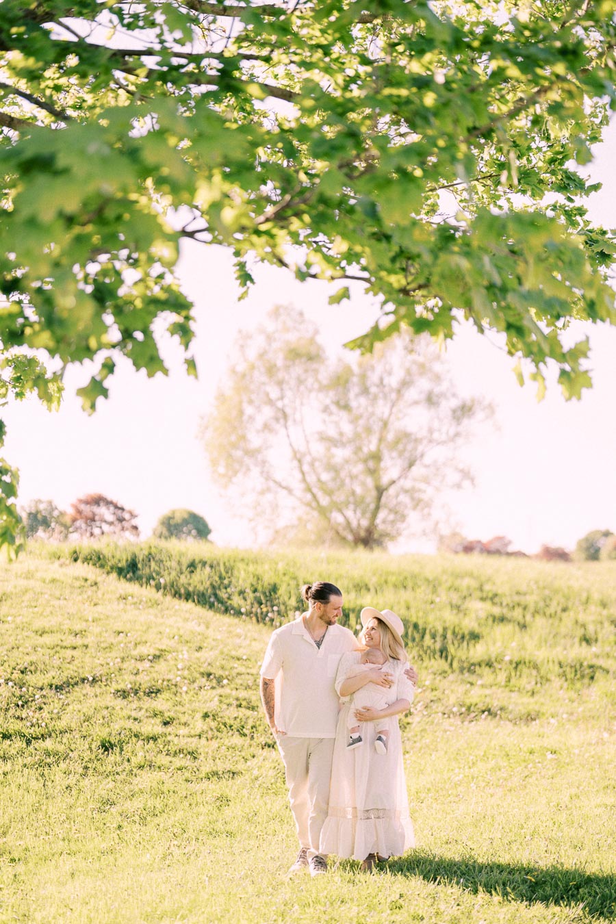 A couple walks through a sunlit park, holding their baby. The scene is serene, with lush green grass and a large tree providing shade. The family appears happy and relaxed, enjoying a peaceful day outdoors.