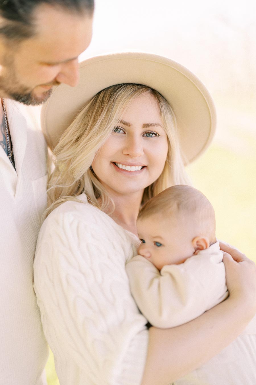 A happy family moment captured outdoors, featuring a woman in a hat holding a baby, with a man beside her.