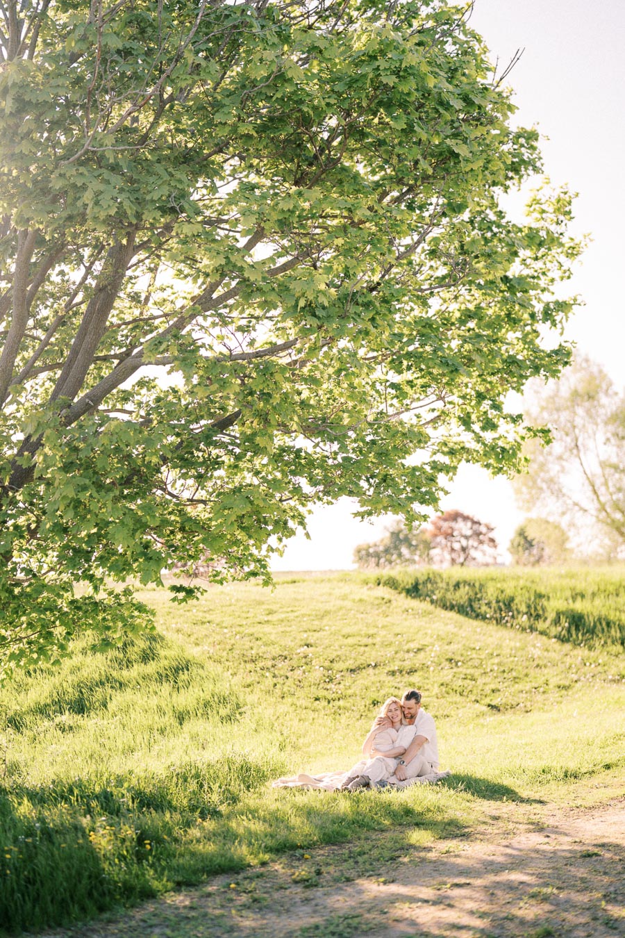 A couple enjoys a peaceful moment sitting on a grassy field under a large tree, basking in the sunlight.