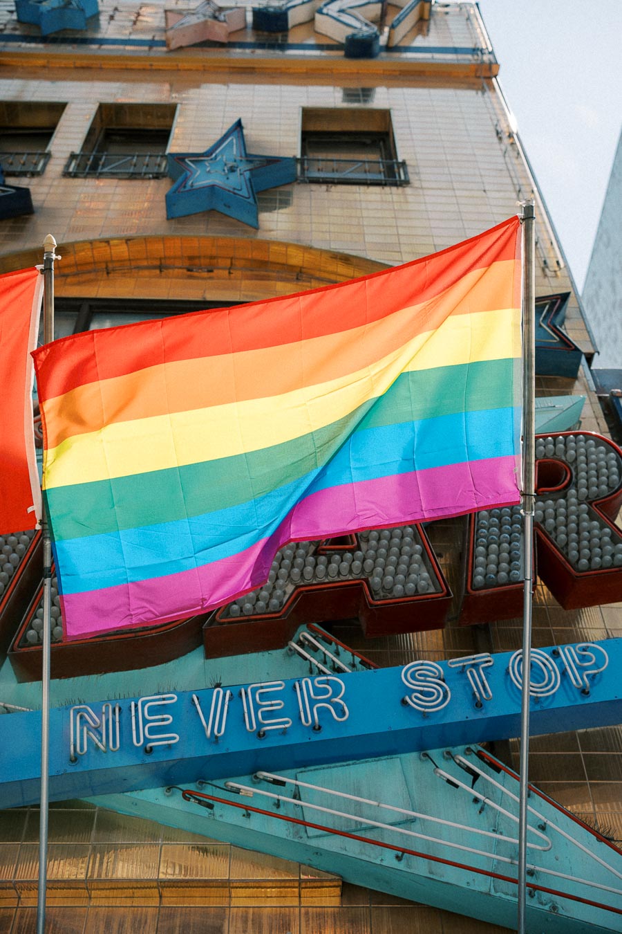 A vibrant rainbow flag waves in front of a building adorned with a neon sign that reads 'NEVER STOP', symbolizing pride and resilience.