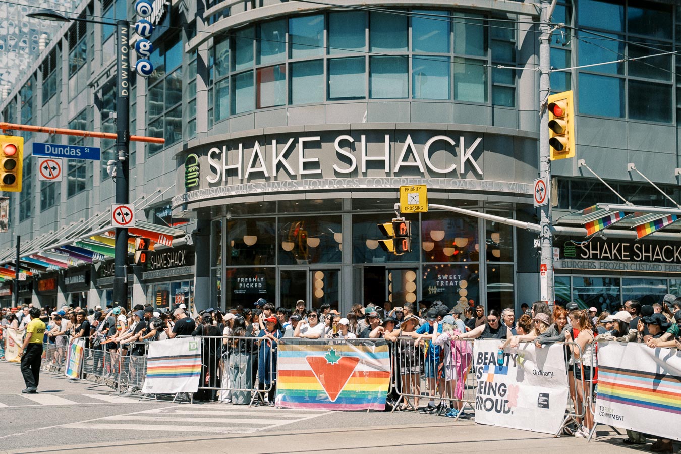 A large crowd gathers outside Shake Shack on Dundas Street, adorned with rainbow banners, likely during a Pride event.