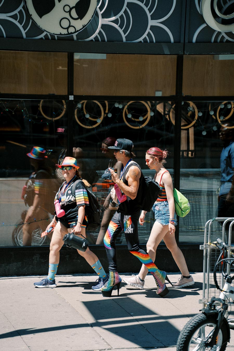 Three individuals in vibrant, rainbow-themed attire walk past a shop window, reflecting their colorful outfits and accessories.