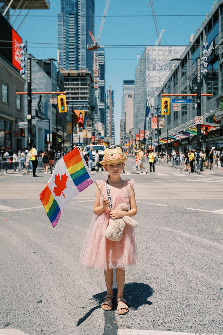 A young girl stands in a city street holding a Canadian flag with rainbow stripes, symbolizing inclusivity and pride.