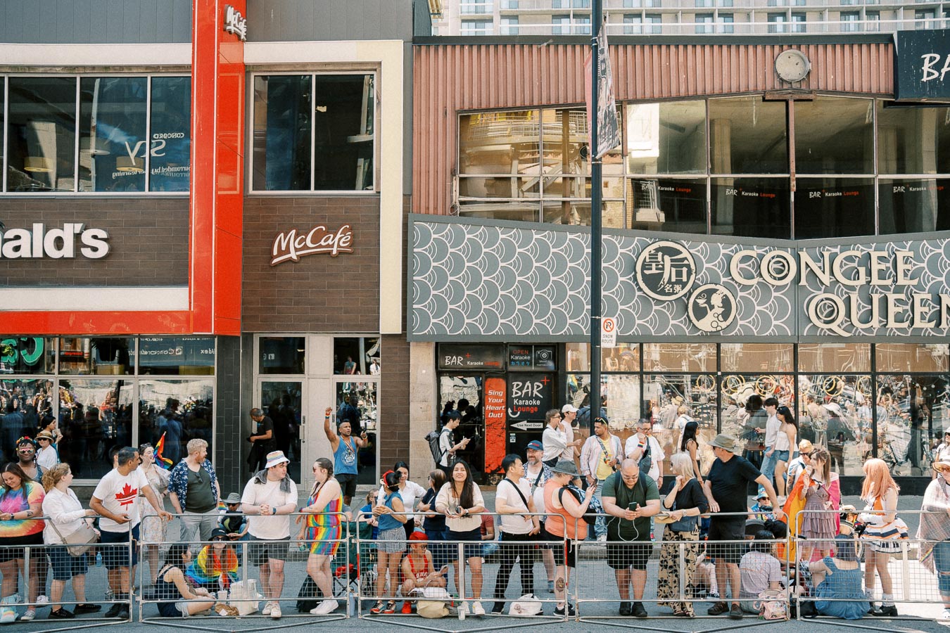 A diverse crowd gathers outside McDonald's and Congee Queen, likely during a pride event, as indicated by the colorful attire and rainbow flags.