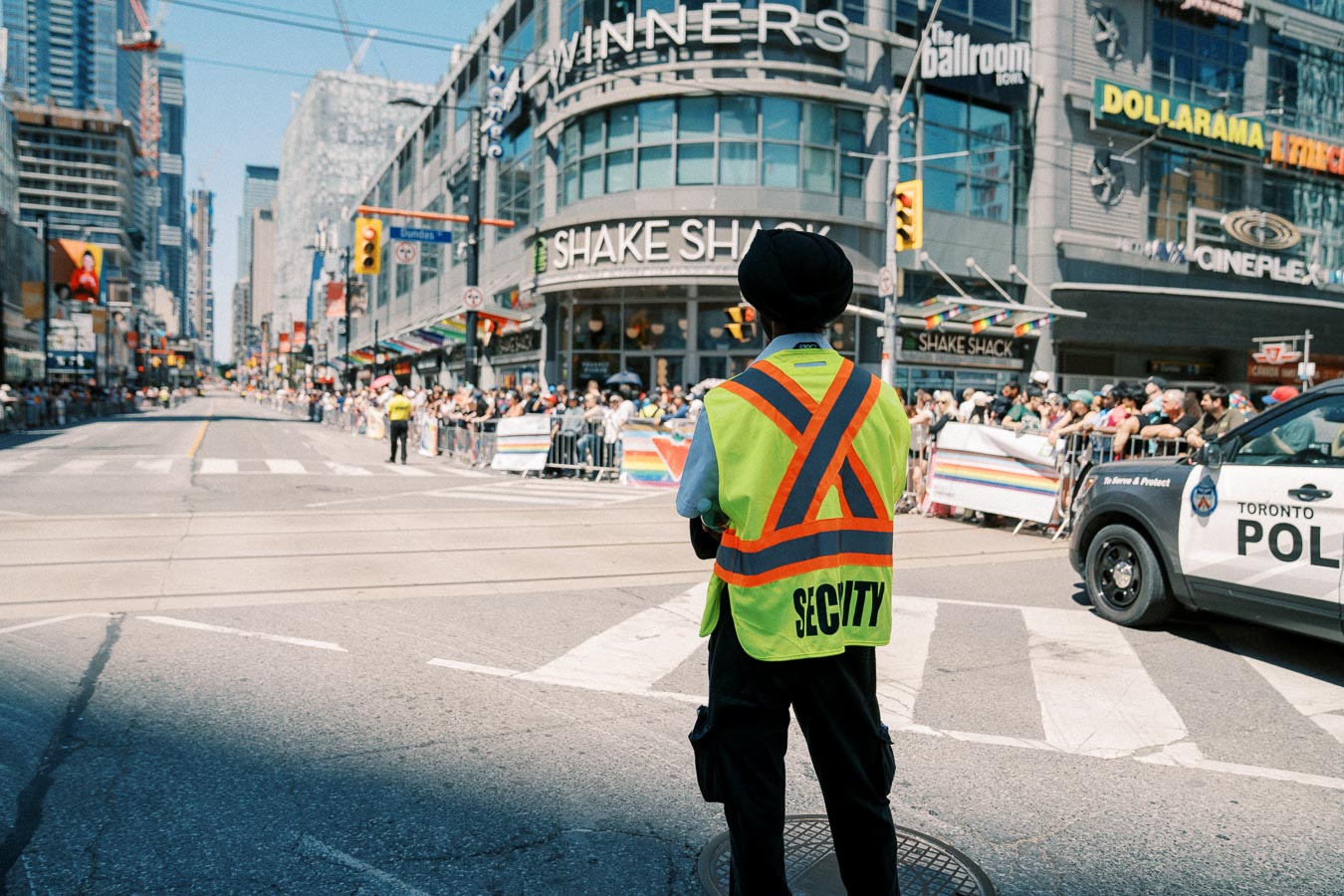 A security guard oversees a parade in Toronto, with a crowd gathered along the street and a police car present.