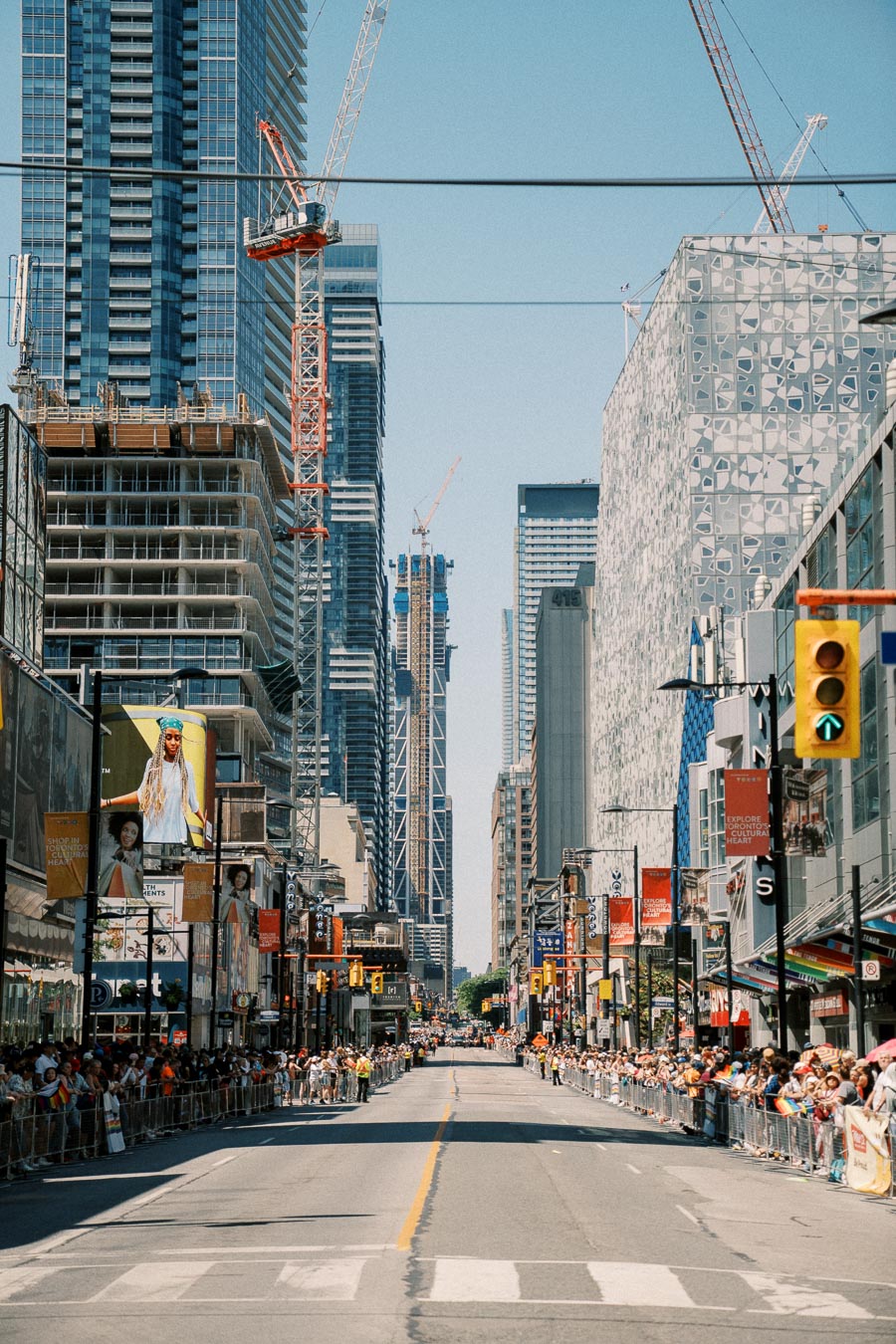 A city street bustling with people lined up on both sides, surrounded by tall skyscrapers and construction cranes, indicating an urban event or parade.