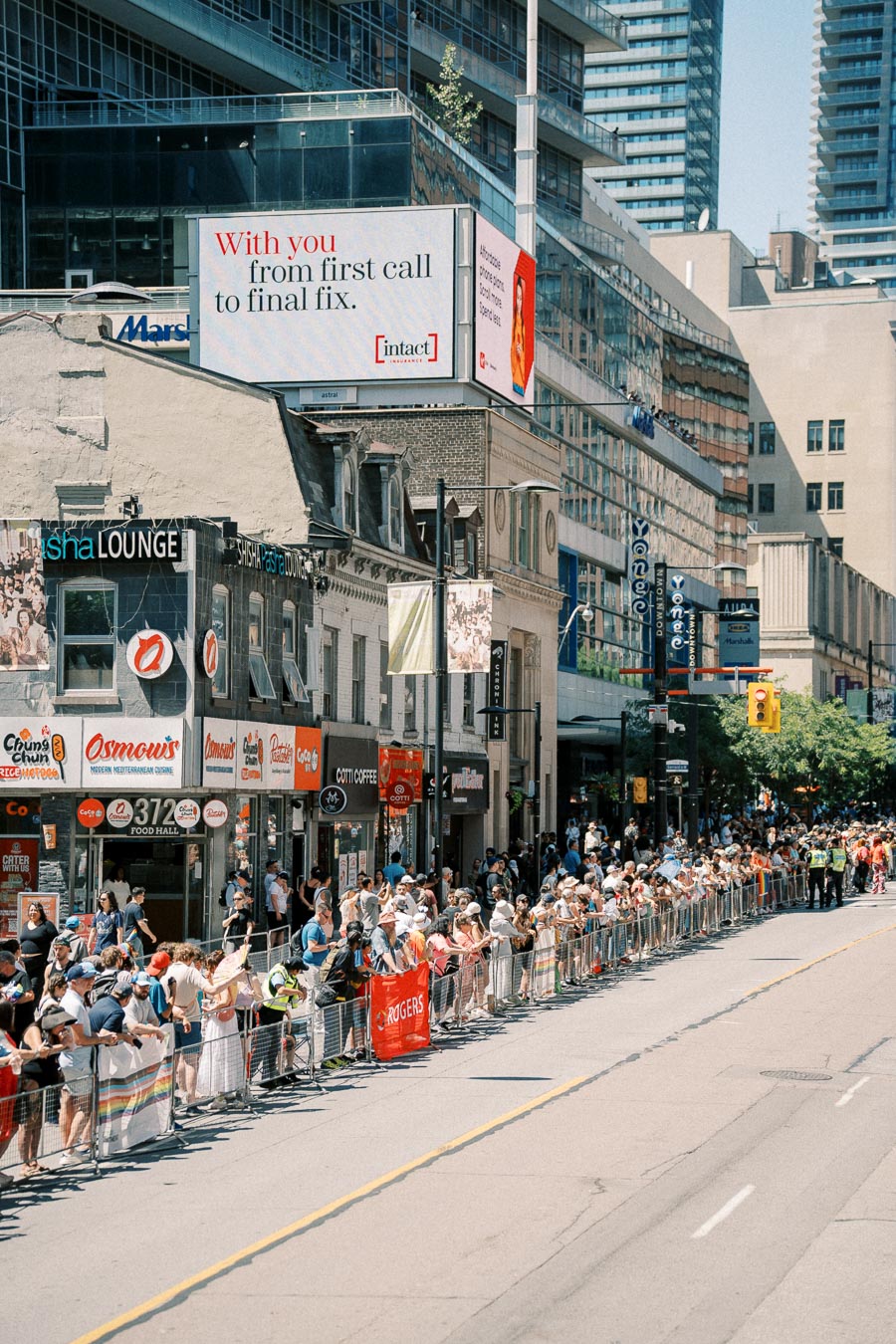 Crowds gather along a city street, possibly for a parade or event, with storefronts and a large billboard in the background.