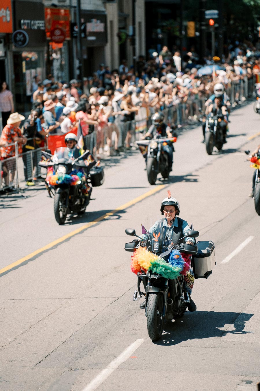 Motorcyclists adorned with rainbow decorations ride in a pride parade, cheered on by a crowd of spectators.