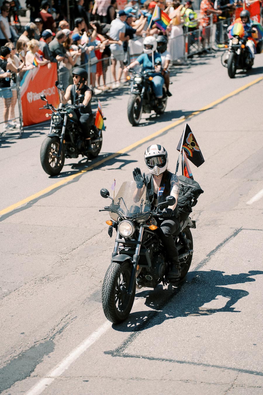 Motorcyclists ride in a parade, waving flags and greeting spectators along the street.