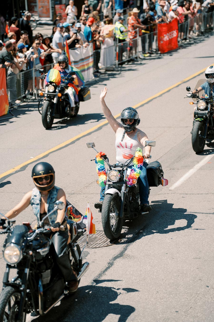Motorcyclists ride in a pride parade, adorned with rainbow decorations and flags, as they wave to the cheering crowd.