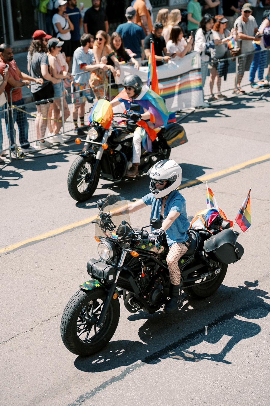 Motorcyclists participate in a pride parade, displaying rainbow flags and decorations, as spectators line the street.