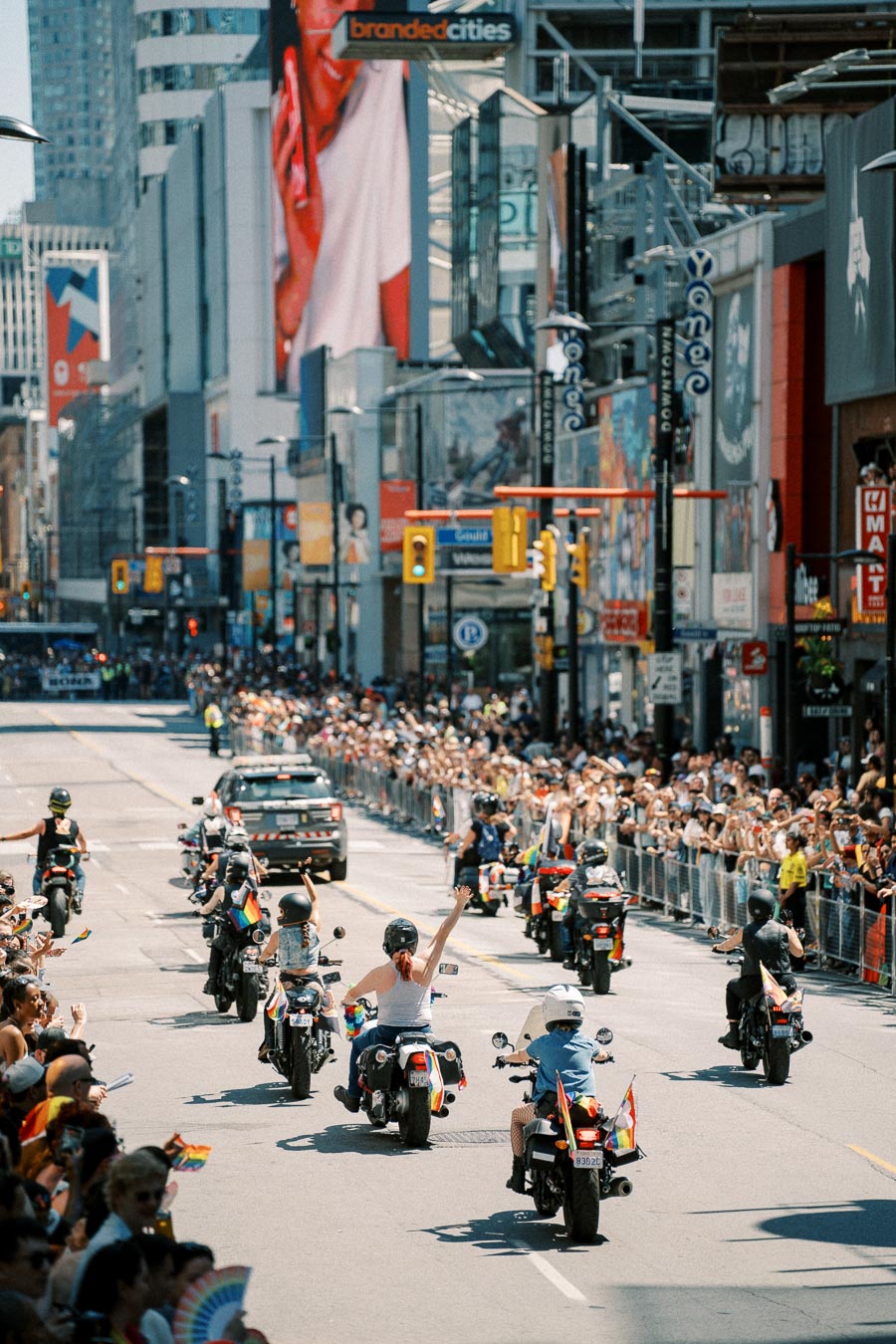 Motorcyclists with rainbow flags participate in a parade on a bustling city street, cheered on by large crowds.