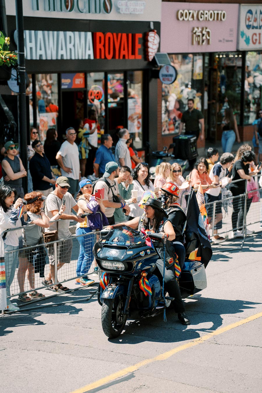 A parade participant rides a motorcycle adorned with rainbow decorations, passing by a crowd of onlookers outside shops.