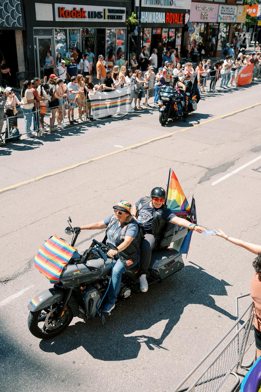 Two individuals ride a motorcycle adorned with rainbow flags during a parade, engaging with spectators lining the street.