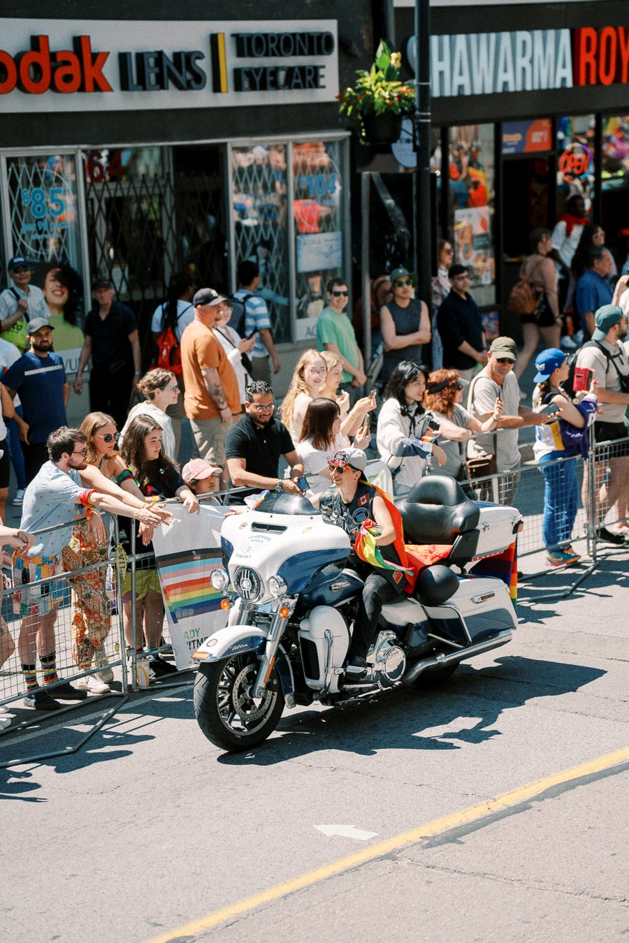 A lively parade scene with a person riding a motorcycle, adorned with rainbow flags, passing by a cheering crowd in front of shops.