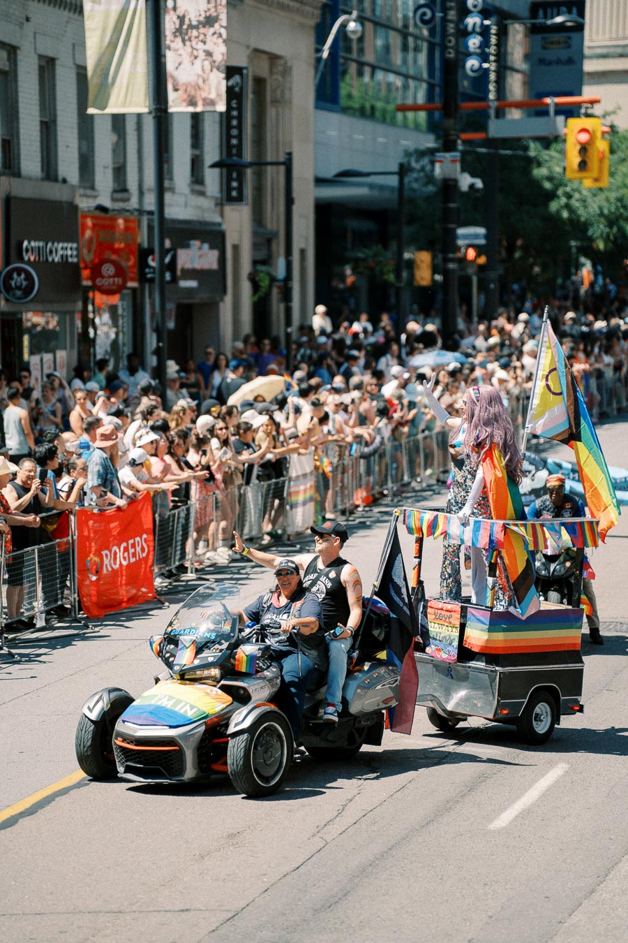 A vibrant parade scene featuring a decorated vehicle with rainbow flags and people waving, surrounded by a cheering crowd on a city street.