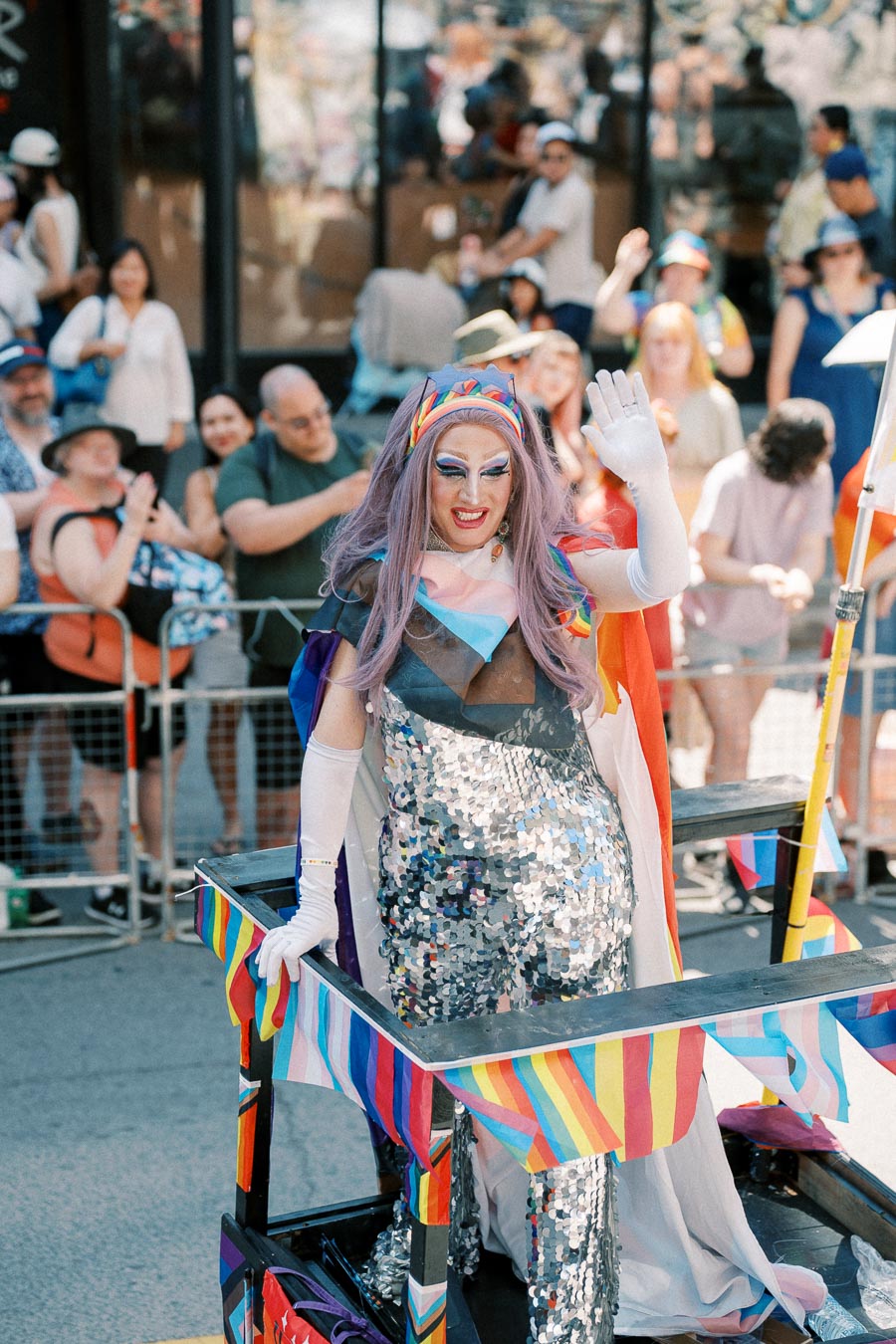 A vibrant drag performer waves to the crowd during a lively parade, adorned in a sequined outfit and rainbow-themed accessories.