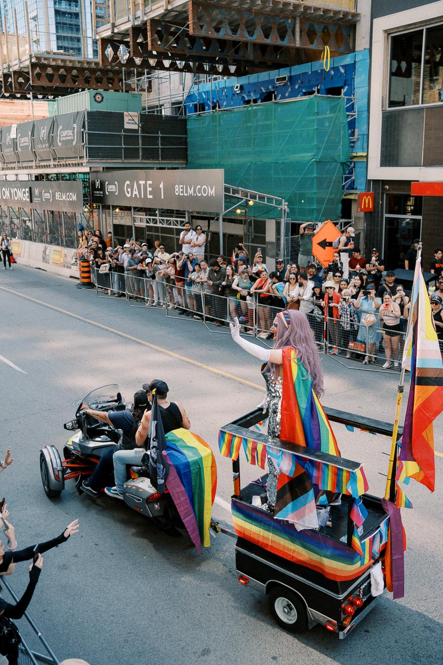 A vibrant parade scene featuring a float adorned with rainbow flags and a person in a colorful outfit waving to the crowd, set against an urban backdrop.