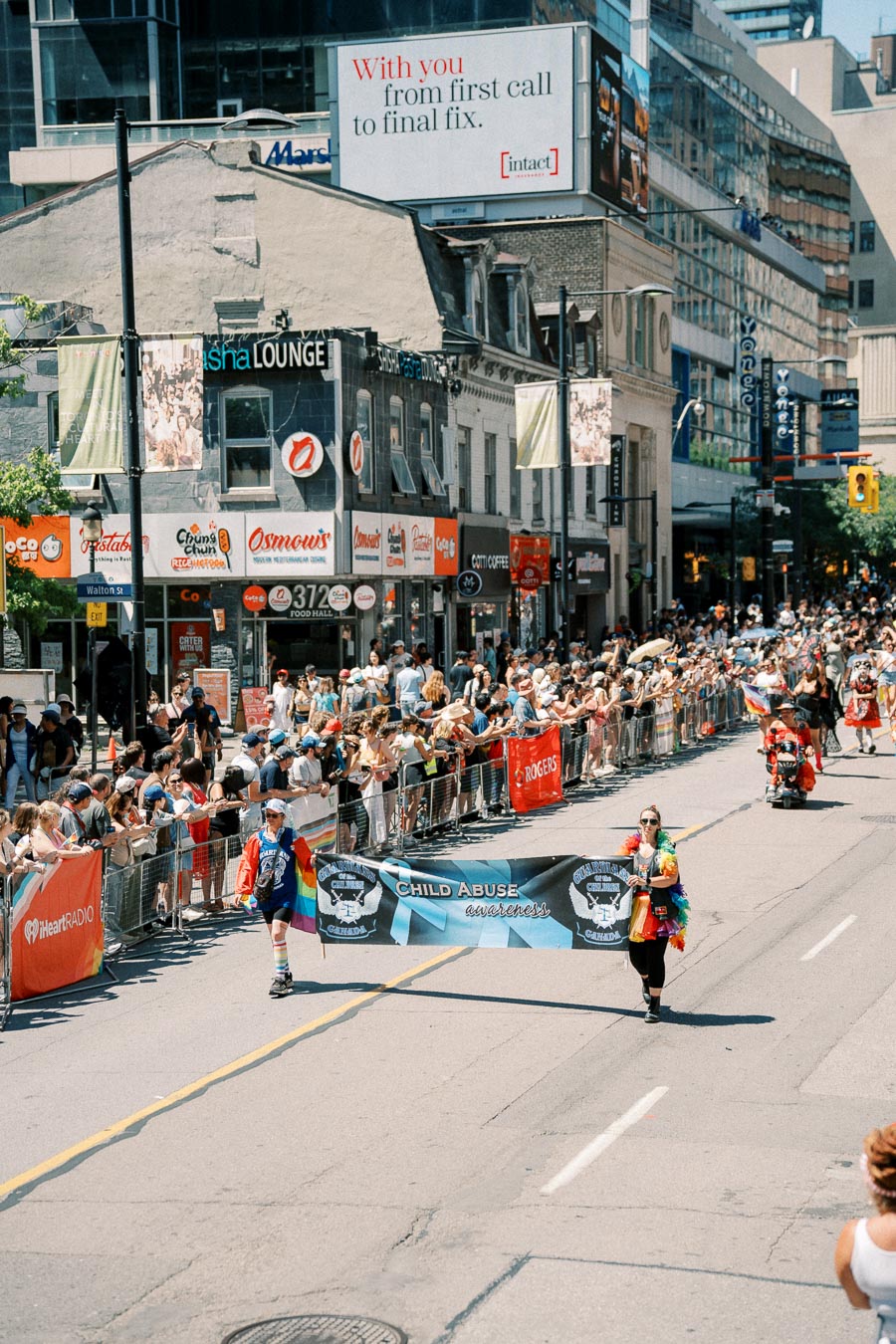 A parade takes place on a busy city street, with participants holding a banner promoting child abuse awareness. Spectators line the sidewalks, and various storefronts and advertisements are visible in the background.