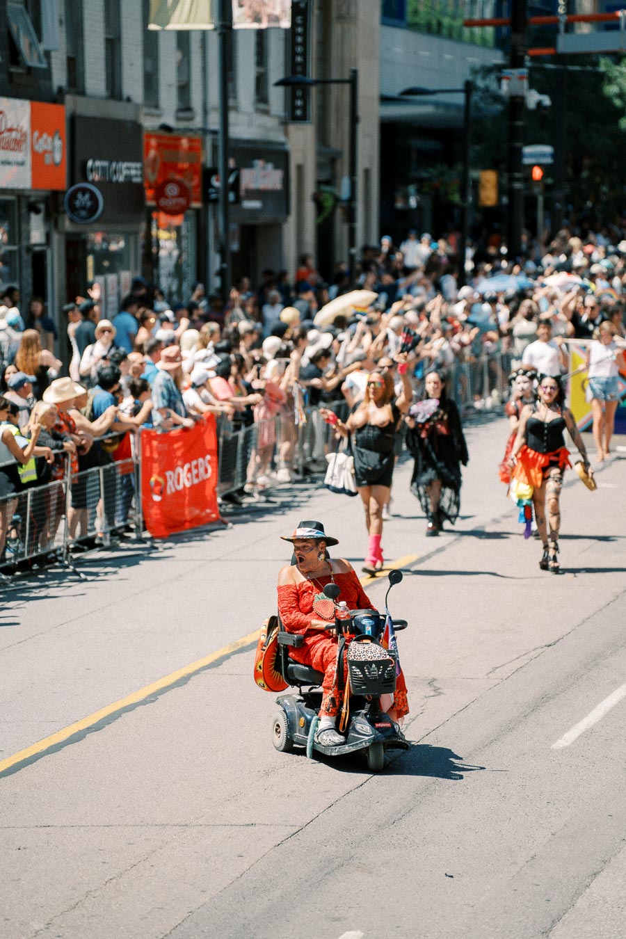 A vibrant parade scene featuring a person in a red outfit riding a mobility scooter, with a lively crowd cheering along the street.
