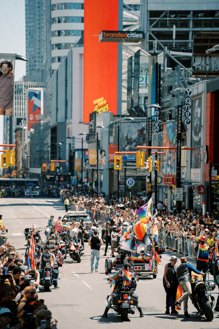 A lively pride parade featuring motorcycles adorned with rainbow flags moves through a bustling city street, surrounded by cheering crowds.