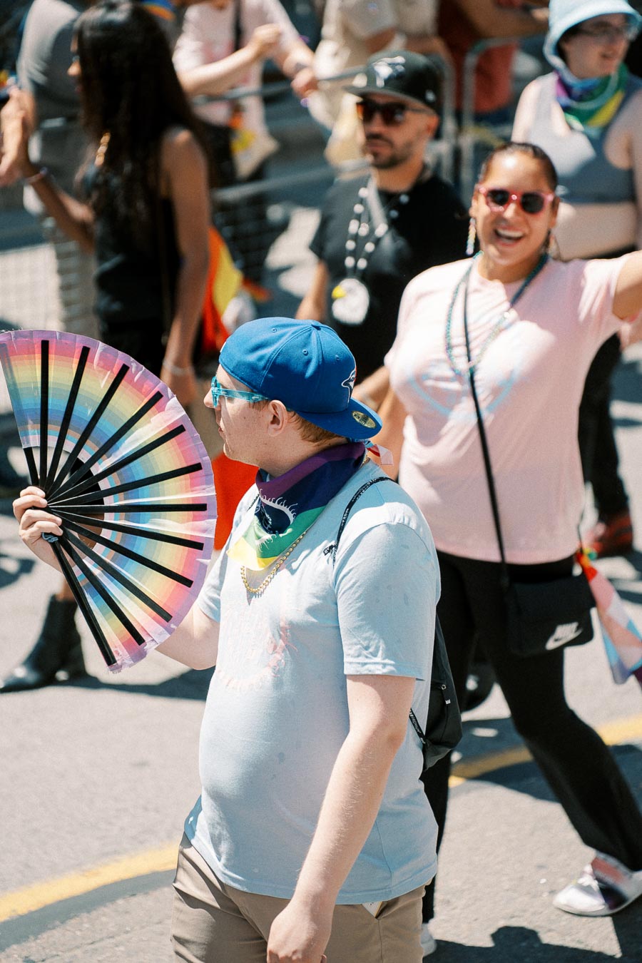 Participants in a pride parade, with one person holding a rainbow fan and wearing a blue cap.