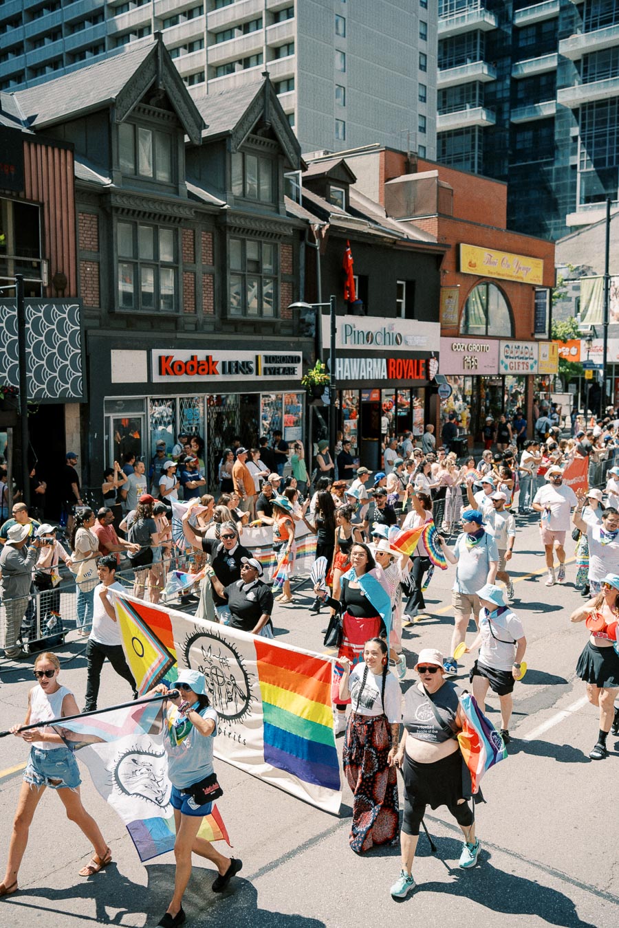 Participants in a colorful parade march down a city street, waving rainbow flags and banners, with spectators lining the sidewalks.