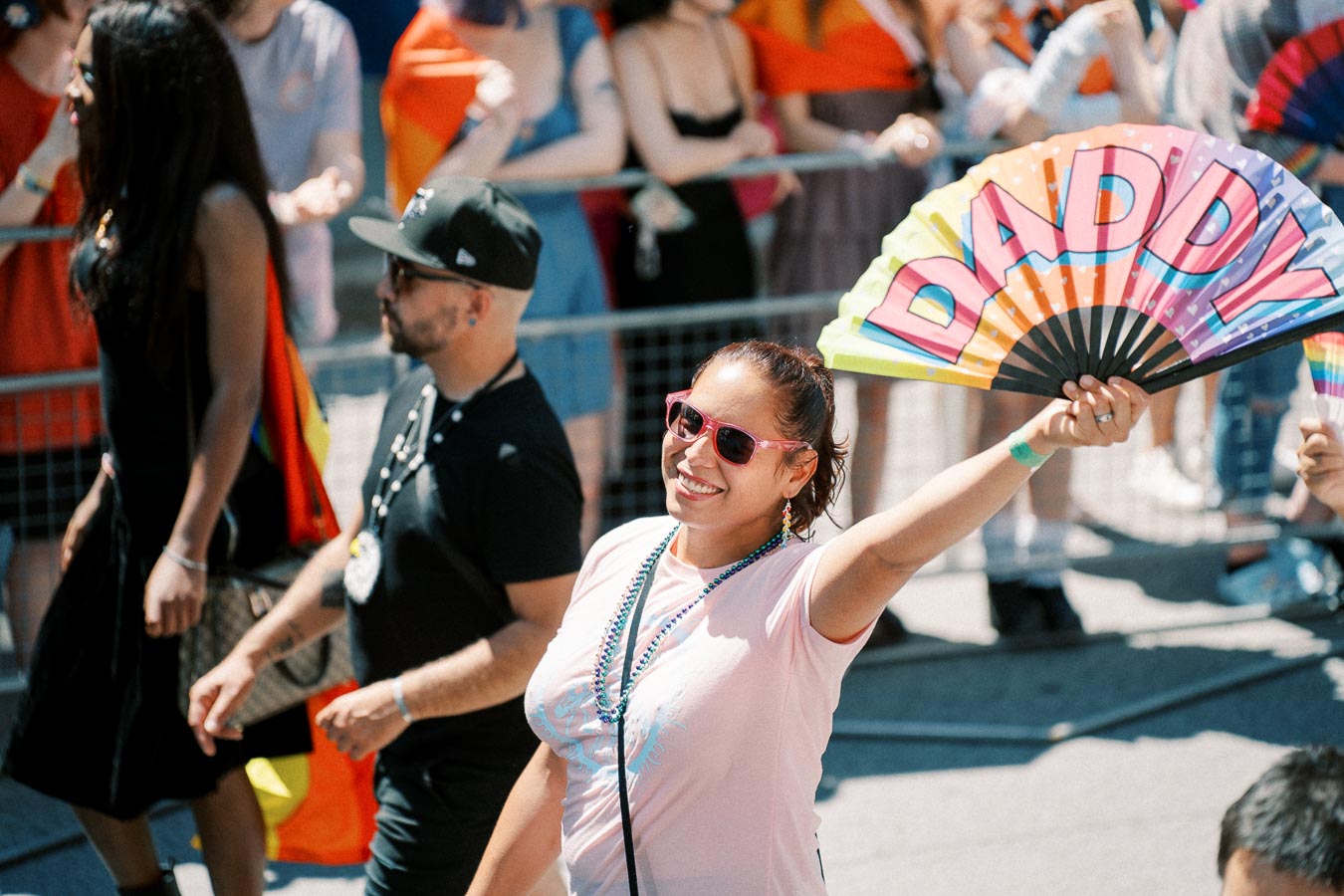 A participant at a pride parade joyfully holds up a colorful fan with the word 'DADDY' written on it, surrounded by a lively crowd.
