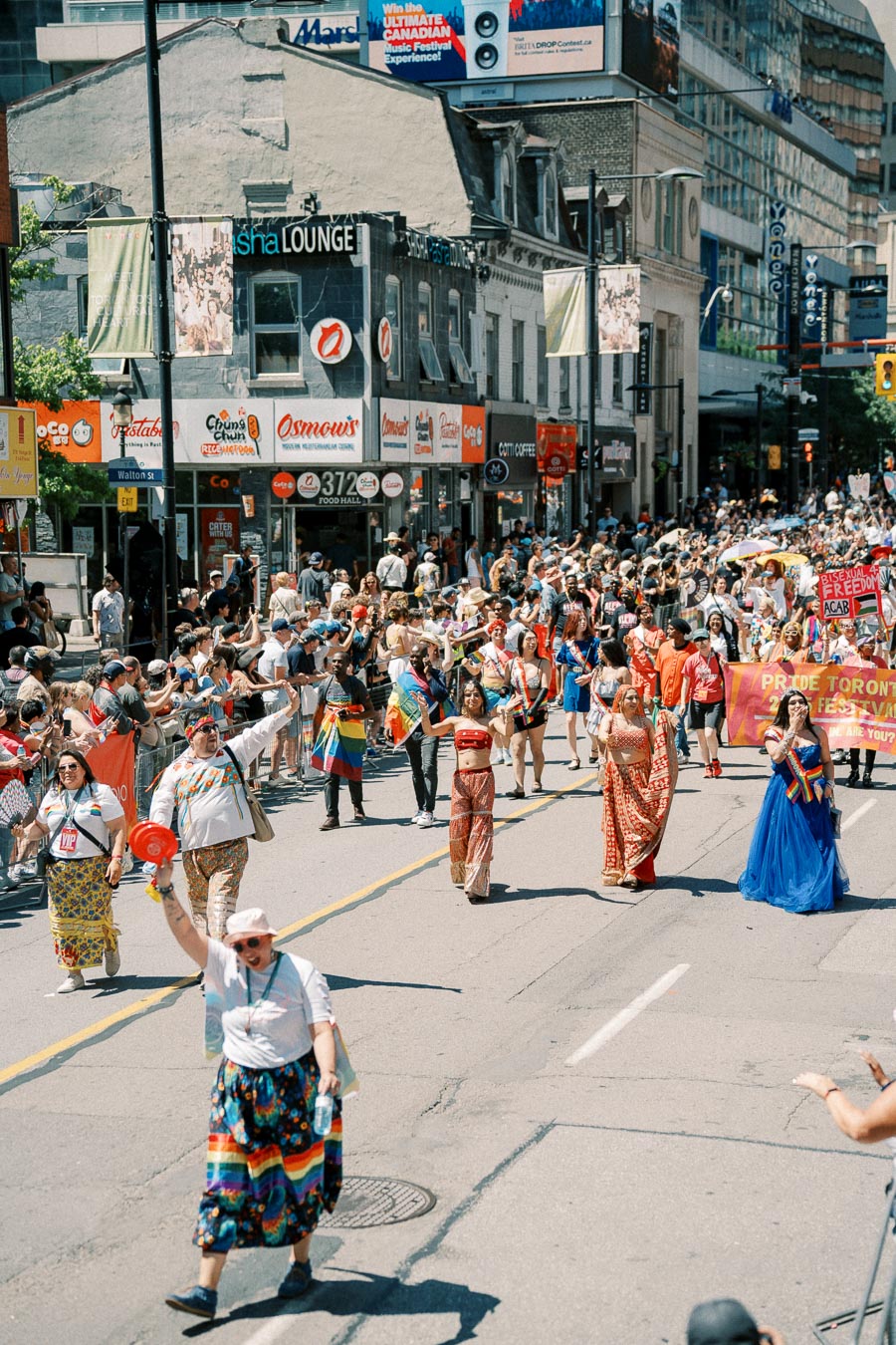 Participants in colorful attire march in a lively parade down a bustling city street, with spectators lining the sidewalks.