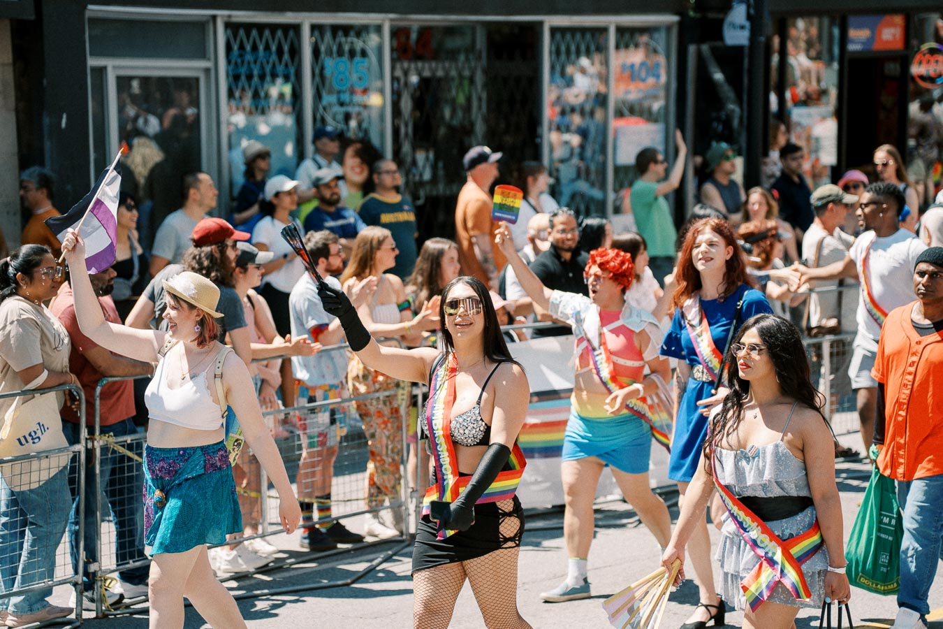 Participants in a vibrant pride parade march down the street, waving flags and wearing colorful attire, while spectators cheer from the sidelines.