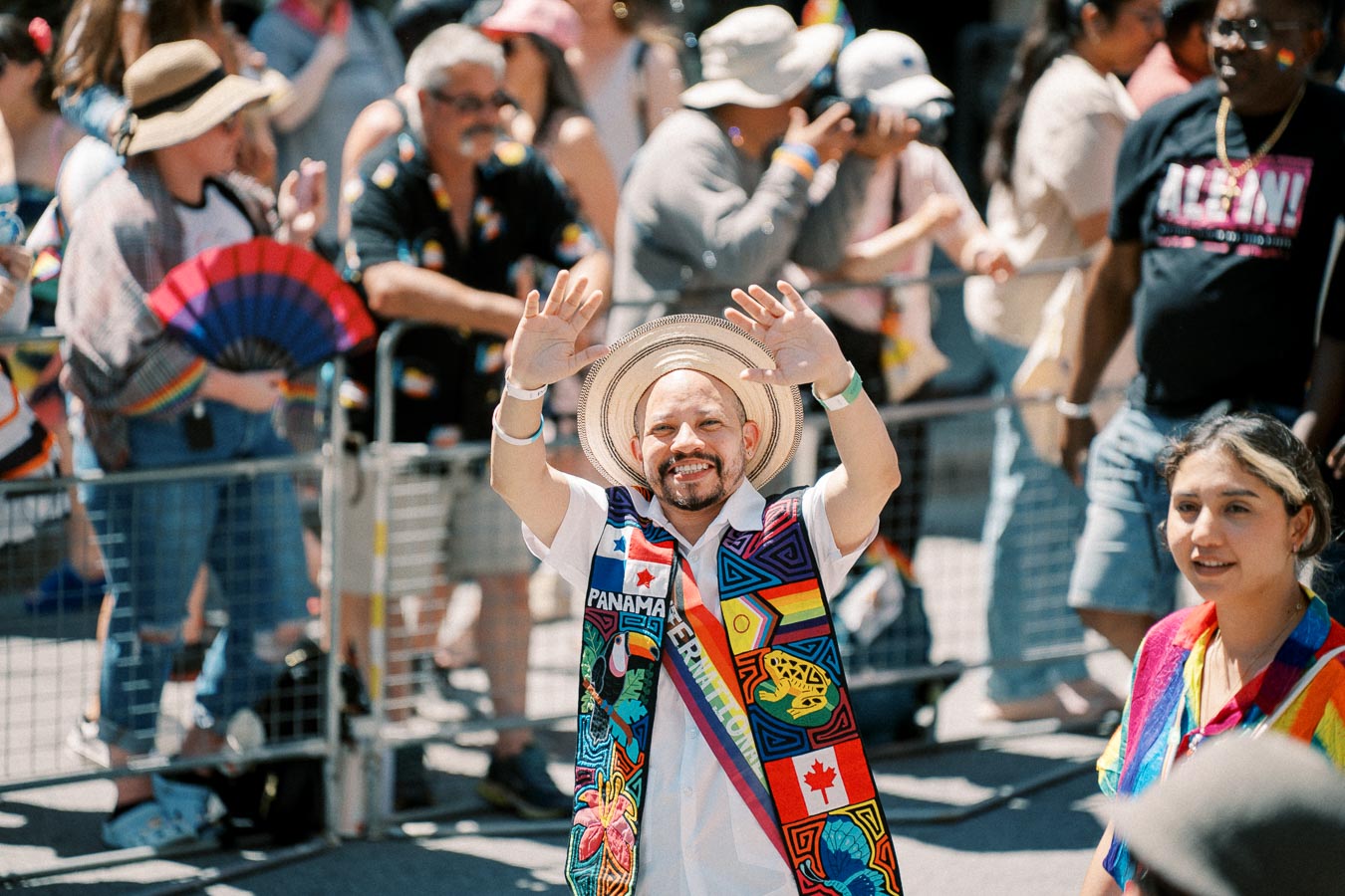 A person in a vibrant traditional outfit and straw hat waves during a lively parade, surrounded by a diverse crowd.