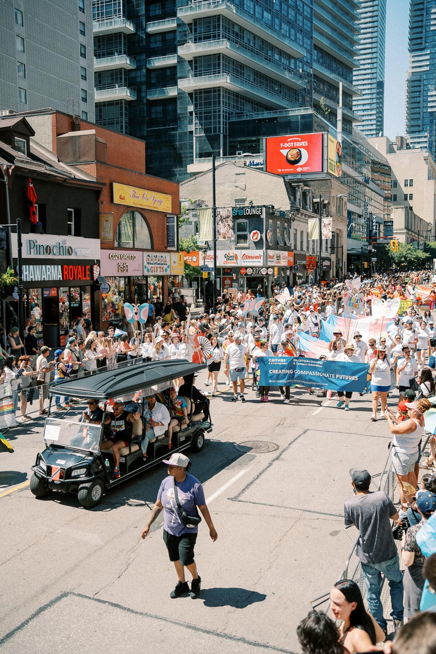 A vibrant parade takes place on a city street, with participants carrying banners and flags. Spectators line the sidewalks, and urban buildings with various storefronts are visible in the background.