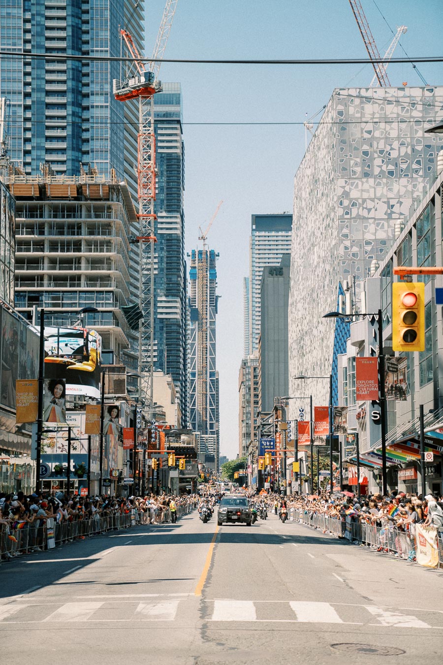 A vibrant city street scene featuring a parade with crowds gathered along the sidewalks. Tall skyscrapers and construction cranes dominate the skyline, indicating ongoing urban development.