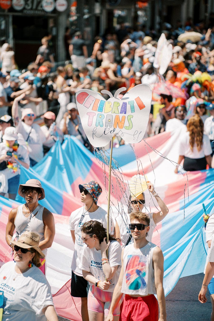 Participants in a pride parade hold a large transgender flag and a sign that reads 'Celebrate Trans Kids'.