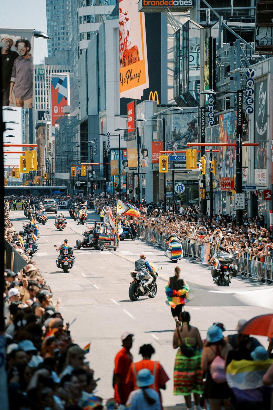 A lively pride parade takes place on a bustling city street, featuring motorcycles and colorful floats adorned with pride flags. Enthusiastic crowds line the sidewalks, celebrating diversity and inclusion.