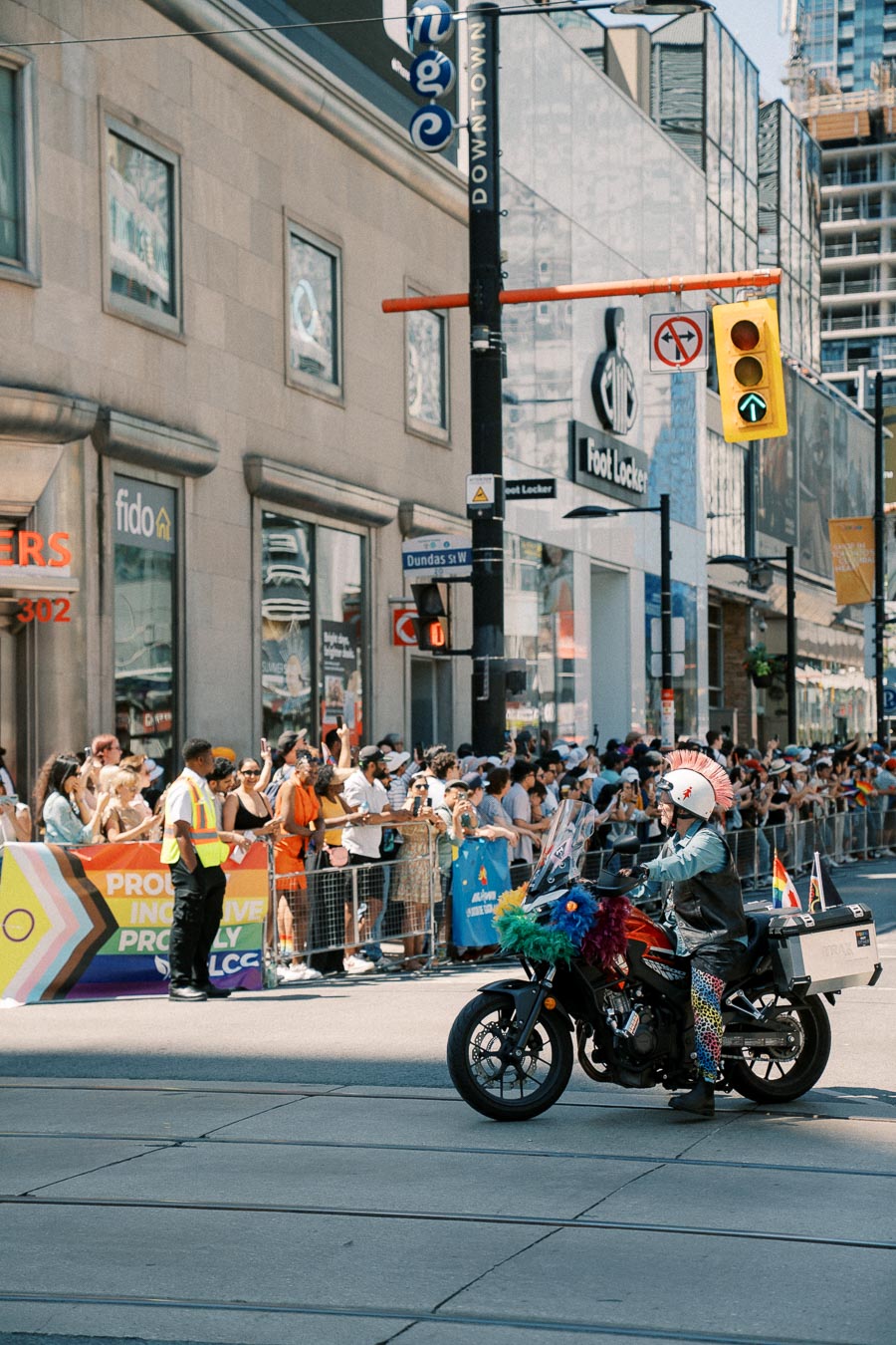 A motorcycle decorated with colorful feathers and flags rides through a parade in an urban area, with a crowd gathered on the sidewalk.