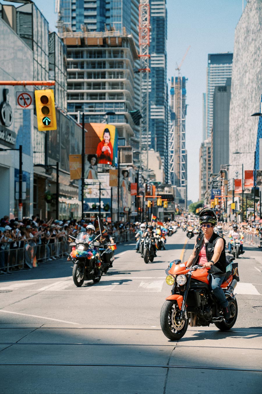 Motorcyclists participate in a city parade, with spectators lining the street and skyscrapers in the background.