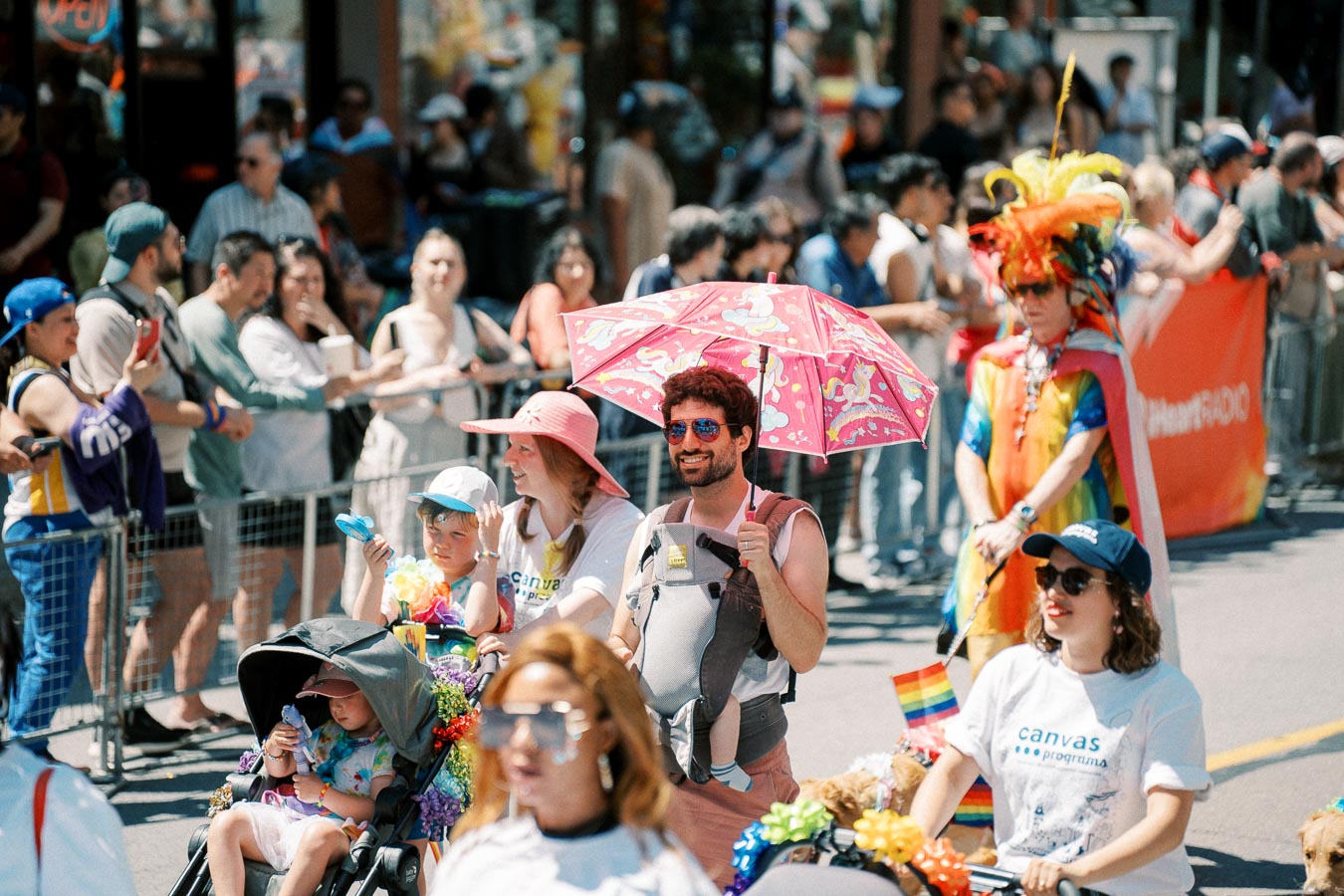Participants in a vibrant parade, featuring colorful outfits and a pink unicorn umbrella, enjoy the festivities as onlookers cheer from the sidelines.