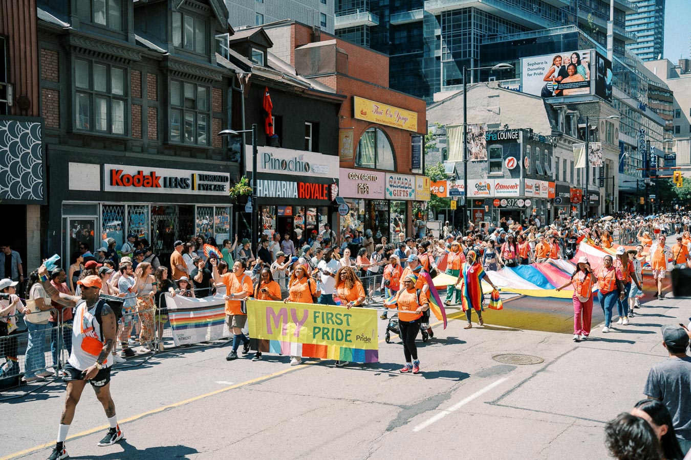 A lively Pride parade takes place on a bustling city street, featuring participants with a 'My First Pride' banner. Spectators line the sidewalks, and storefronts like Kodak Lens and Pinocchio are visible.