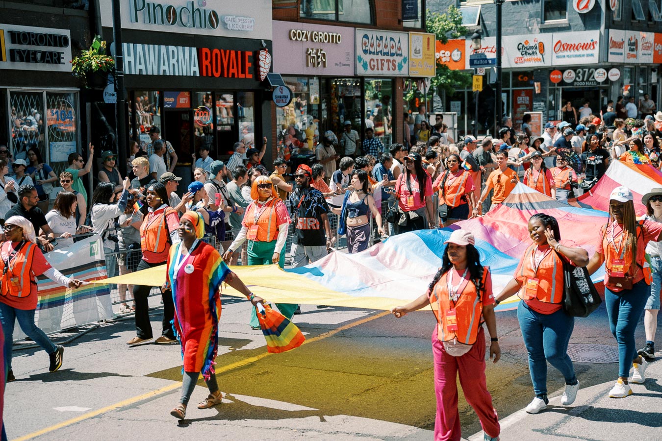 Participants in a colorful parade carry a large rainbow flag down a bustling street, surrounded by cheering spectators and vibrant storefronts.