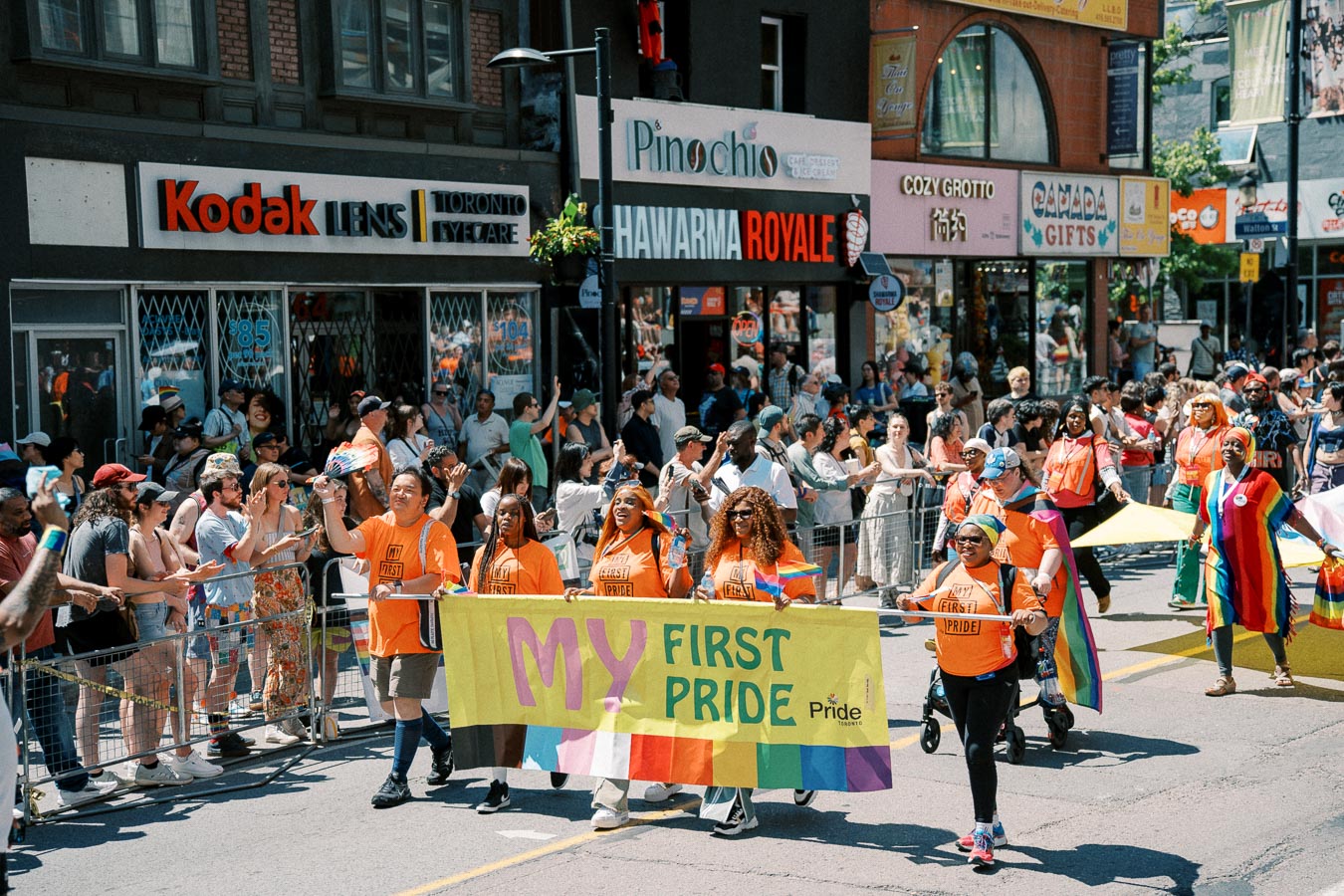Participants in a Pride parade march with a 'My First Pride' banner, celebrating diversity and inclusion. Spectators line the street, and shops are visible in the background.