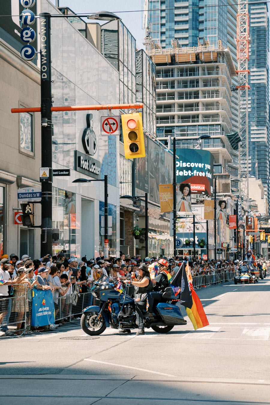 A vibrant parade takes place on a bustling city street, featuring motorcycles adorned with flags. Spectators line the sidewalks, and the scene is set against a backdrop of tall buildings, including a Foot Locker store.