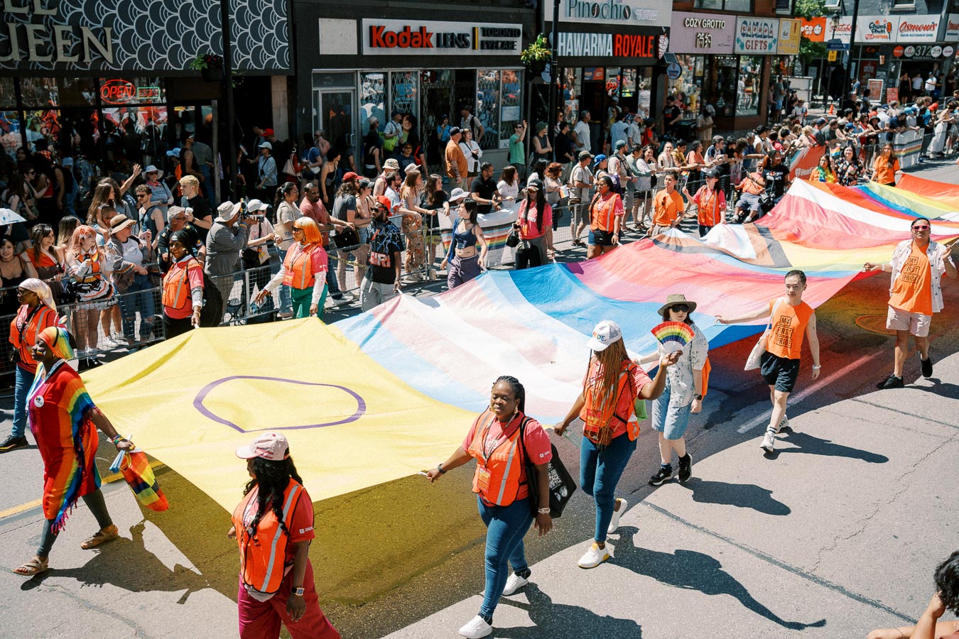 Participants in a parade carry a large rainbow flag, celebrating diversity and inclusion, as onlookers cheer from the sidelines.