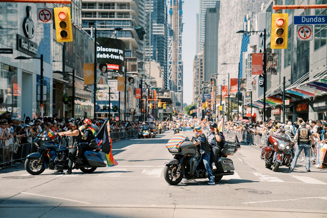 Motorcycle riders with rainbow flags participate in a pride parade on a busy city street, surrounded by spectators.